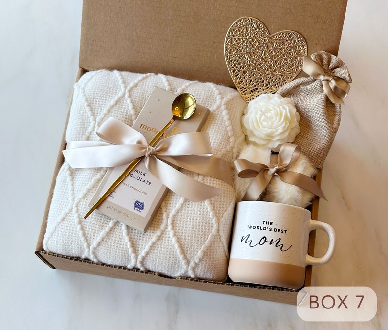 May include: A gift box filled with a white blanket, a bar of milk chocolate, a gold spoon, a white flower, a small bag, a heart-shaped gold ornament, and a white mug with the text "The World's Best Mom" in black. The box is labeled "Box 7".