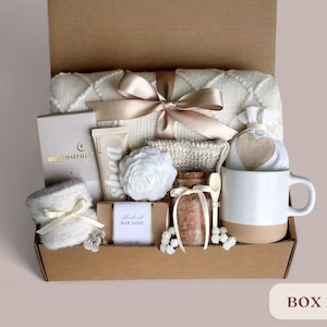 May include: A brown cardboard box filled with a variety of self-care items, including a white throw blanket, a white mug, a bar of soap, a jar of bath salts, a white hand towel, a white hand cream, a white bath puff, and a white and brown heart-shaped pouch. The box is labeled "BOX 1".