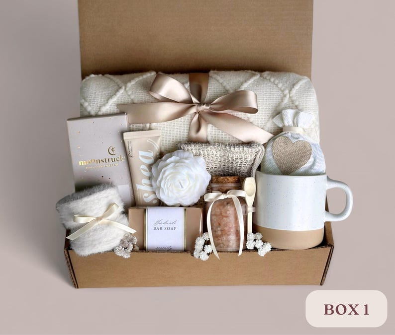 May include: A brown cardboard box filled with a variety of self-care items, including a white throw blanket, a white mug, a bar of soap, a jar of bath salts, a white hand towel, a white hand cream, a white bath puff, and a white and brown heart-shaped pouch. The box is labeled "BOX 1".