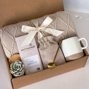 K&ouml;nnte beinhalten: Eine Geschenkbox gef&uuml;llt mit gem&uuml;tlichen Artikeln, darunter eine beige gestrickte Wolldecke, eine Tasse, eine kleine Tasche, ein goldener L&ouml;ffel, eine Sukkulente in einem Topf und eine Tafel Milchschokolade mit dem Text "moonstruck" auf der Verpackung.
