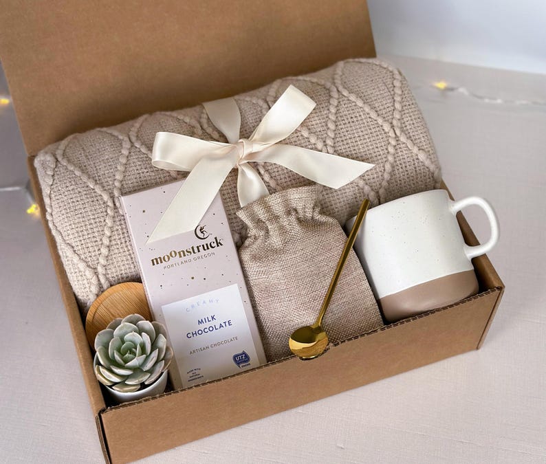 K&ouml;nnte beinhalten: Eine Geschenkbox gef&uuml;llt mit gem&uuml;tlichen Artikeln, darunter eine beige gestrickte Wolldecke, eine Tasse, eine kleine Tasche, ein goldener L&ouml;ffel, eine Sukkulente in einem Topf und eine Tafel Milchschokolade mit dem Text "moonstruck" auf der Verpackung.