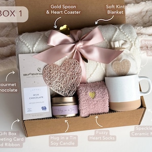 Peut inclure: Une bo&icirc;te-cadeau remplie d'articles de soins personnels, comprenant une couverture en tricot blanc doux, une chaussette en forme de c&oelig;ur rose et moelleuse, une tasse en c&eacute;ramique mouchet&eacute;e, une barre de chocolat gastronomique, une bougie de soja et une boisson dans un sac en toile de jute. La bo&icirc;te est &eacute;tiquet&eacute;e "BOX 1".