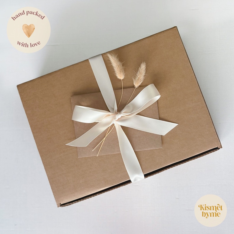 May include: A brown cardboard box with a white ribbon and dried flowers. The box is tied with a white ribbon and has a tag that says "hand packed with love".