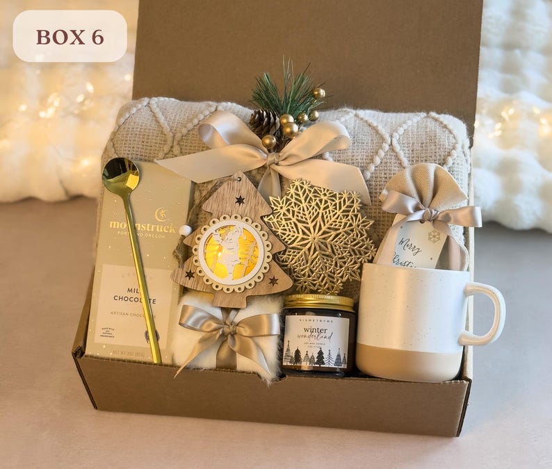 May include: A brown gift box filled with a cozy winter-themed gift set. The box contains a white blanket, a gold spoon, a milk chocolate bar, a wooden Christmas tree ornament, a snowflake ornament, a jar of winter wonderland scented candle, a small white mug with a brown band, and a small burlap bag with the words "Merry & Bright" written on it.
