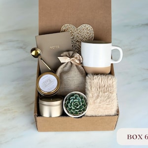 May include: A brown cardboard box filled with a cozy gift set. The set includes a white mug with a beige bottom, a small succulent plant in a white pot, a candle, a small bag of brown burlap, a gold spoon, a notebook with the word "NOTES" on the cover, and a fluffy white throw. The box is labeled "BOX 6".