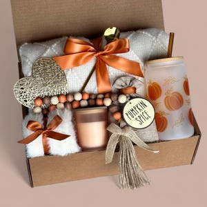 May include: A brown cardboard gift box filled with fall-themed items, including a white blanket, a candle, a heart-shaped ornament, a wooden bead garland, a burlap bag with a tassel, a tumbler with a pumpkin spice design, and a wooden tag that says "Pumpkin Spice".