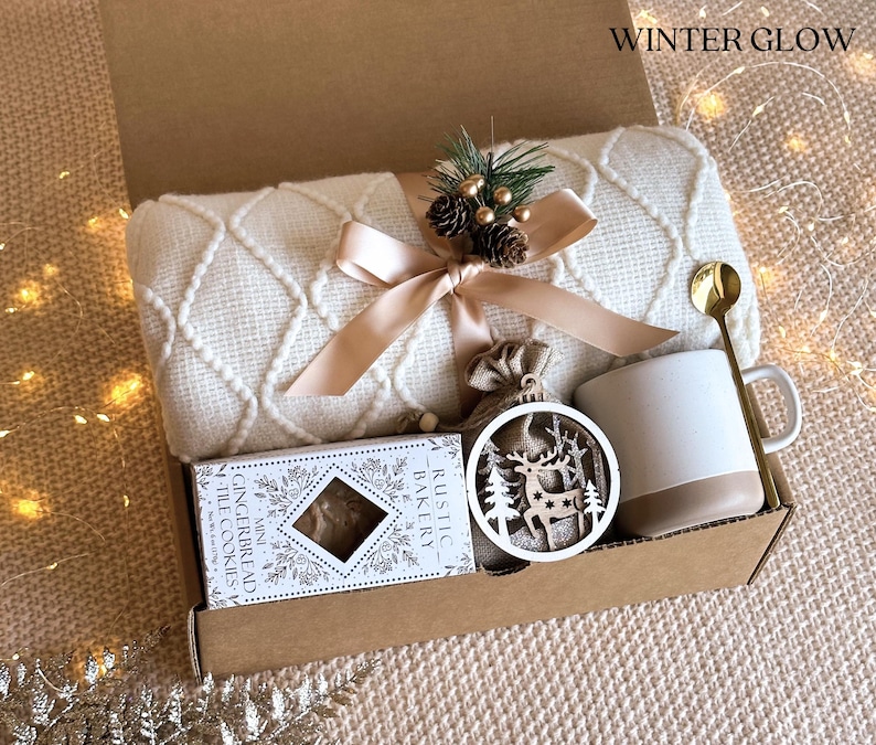 May include: A gift box filled with cozy winter items, including a white knitted throw blanket, a mug, a wooden reindeer ornament, a bag of cookies, and a gold spoon. The box is labeled "Rustic Bakery" and the ornament says "Winter Glow".