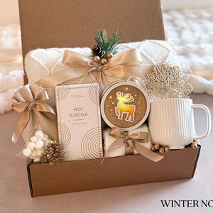 Puede incluir: Una caja de regalo con tem&aacute;tica invernal. Incluye una manta blanca, chocolate caliente, una taza, un adorno de madera con dise&ntilde;o de reno y lazos decorativos. La caja es marr&oacute;n y los art&iacute;culos est&aacute;n dispuestos para un ambiente acogedor y festivo. El texto "WINTER NOEL" est&aacute; abajo.