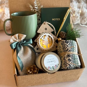 Op de afbeelding: Een bruine cadeaudoos gevuld met een groene mok, een jutezak met een groen lint, een houten kerstboomversiering, een kaars met een gouden deksel en een wit etiket met de tekst "Winter Cranberry", en een paar groen-witte gestreepte sokken.