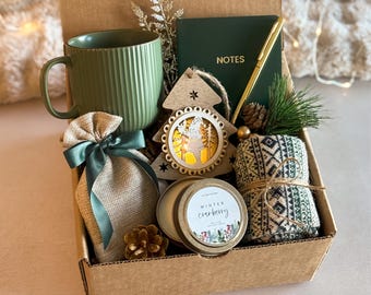 May include: A brown gift box filled with a green mug, a burlap bag with a green ribbon, a wooden Christmas tree ornament, a candle with a gold lid and a white label that says "Winter Cranberry", and a pair of green and white patterned socks.