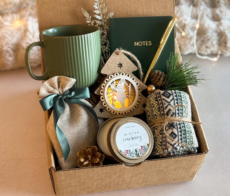 Op de afbeelding: Een bruine cadeaudoos gevuld met een groene mok, een jutezak met een groen lint, een houten kerstboomversiering, een kaars met een gouden deksel en een wit etiket met de tekst "Winter Cranberry", en een paar groen-witte gestreepte sokken.