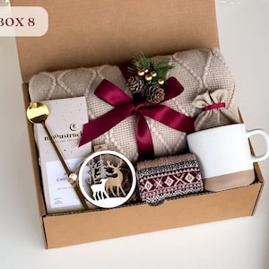 May include: A brown cardboard box with the text "BOX 8" on the top. The box is filled with a variety of gifts, including a beige knitted blanket, a mug, a bag of coffee, a wooden reindeer ornament, and a bar of chocolate.