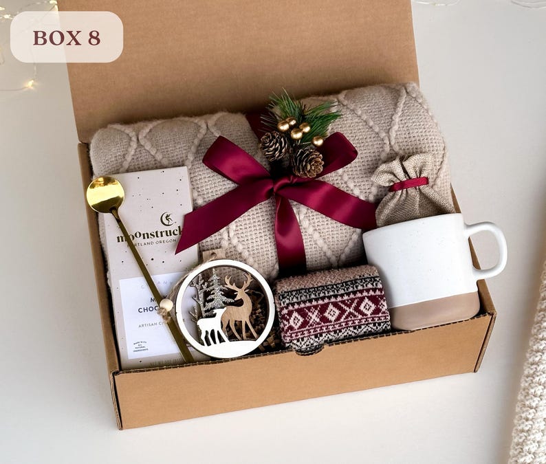 May include: A brown cardboard box with the text "BOX 8" on the top. The box is filled with a variety of gifts, including a beige knitted blanket, a mug, a bag of coffee, a wooden reindeer ornament, and a bar of chocolate.