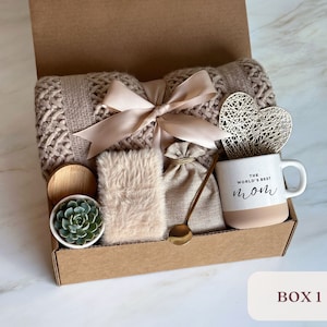 May include: A gift box filled with items for a cozy night in. The box includes a knitted throw blanket, a mug that says "The World's Best Mom", a small succulent plant, a faux fur throw pillow, a small burlap bag, and a gold spoon.