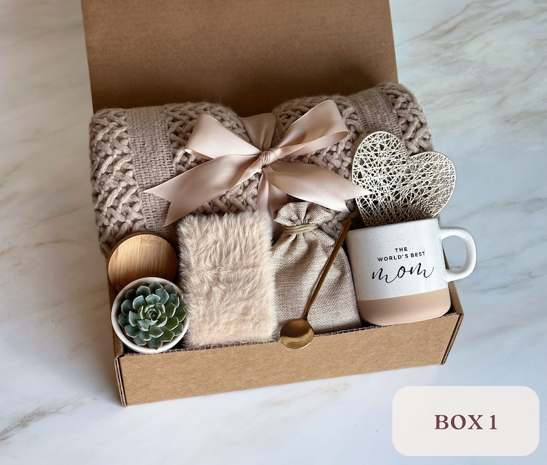 May include: A gift box filled with items for a cozy night in. The box includes a knitted throw blanket, a mug that says "The World's Best Mom", a small succulent plant, a faux fur throw pillow, a small burlap bag, and a gold spoon.
