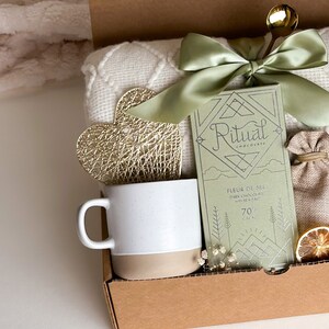 May include: A gift box filled with a white blanket, a mug, a gold heart, a bag of chocolate, a small burlap bag, and dried citrus slices. The chocolate bar is labeled "Ritual Chocolate Fleur de Sel Dark Chocolate with Sea Salt 70% Cacao".
