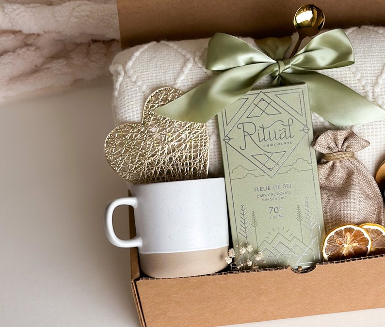 May include: A gift box filled with a white blanket, a mug, a gold heart, a bag of chocolate, a small burlap bag, and dried citrus slices. The chocolate bar is labeled "Ritual Chocolate Fleur de Sel Dark Chocolate with Sea Salt 70% Cacao".