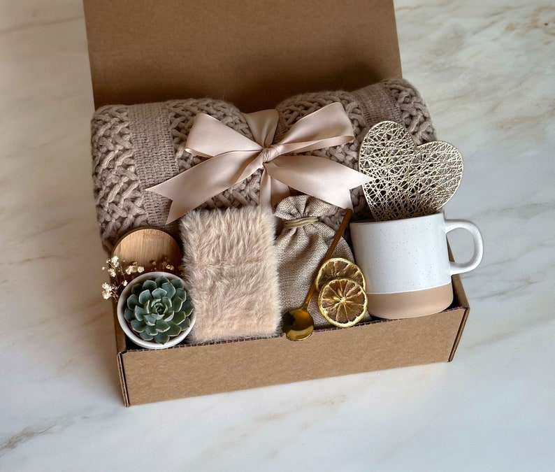 May include: A brown cardboard gift box filled with a variety of items, including a knitted throw blanket, a small potted succulent, a mug, a heart-shaped decorative object, and a small bag. The box is tied with a beige ribbon.