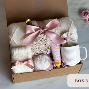 May include: A gift box filled with items for self-care and relaxation. The box includes a white blanket, a mug, a heart-shaped decorative piece, a tube of hand cream, a small white flower, and a small white fluffy item. The box is decorated with a pink ribbon and a bow.