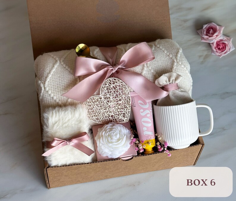 May include: A gift box filled with items for self-care and relaxation. The box includes a white blanket, a mug, a heart-shaped decorative piece, a tube of hand cream, a small white flower, and a small white fluffy item. The box is decorated with a pink ribbon and a bow.