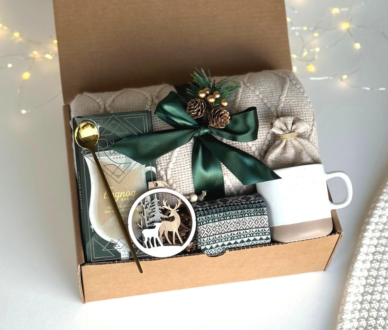 Puede incluir: Una caja de regalo navide&ntilde;a con una manta color crema, una taza blanca y beige, una cuchara dorada, un adorno de ciervo de madera, una caja con estampado verde y blanco y un lazo verde. La caja tambi&eacute;n contiene una tableta de chocolate y una peque&ntilde;a bolsa de yute.