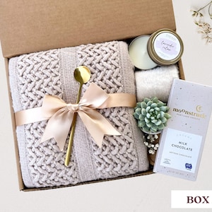 May include: A gift box filled with a cozy knitted throw blanket, a gold spoon, a candle, a succulent plant, and a bar of milk chocolate. The box is tied with a beige ribbon and the chocolate bar has the text "moonstruck" and "creamy milk chocolate" on it.