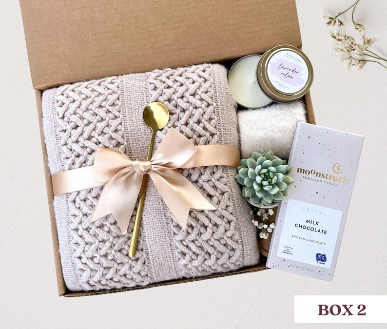 May include: A gift box filled with a cozy knitted throw blanket, a gold spoon, a candle, a succulent plant, and a bar of milk chocolate. The box is tied with a beige ribbon and the chocolate bar has the text "moonstruck" and "creamy milk chocolate" on it.