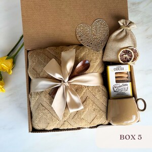 May include: A gift box filled with a brown throw blanket, a wooden spoon, a bag of lemon cookies, a small bag of dried lemon slices, and a brown mug. The box is decorated with a heart-shaped cutout and a ribbon.