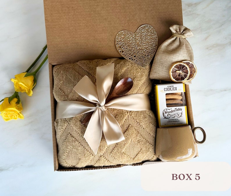 May include: A gift box filled with a brown throw blanket, a wooden spoon, a bag of lemon cookies, a small bag of dried lemon slices, and a brown mug. The box is decorated with a heart-shaped cutout and a ribbon.