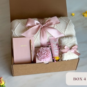 May include: A gift box filled with a white knitted throw blanket, a pink notebook with a gold pen, a pink rose soap, a white fluffy hand towel with a pink ribbon, and a pink hand cream tube with the word "rose" on it. The box is labeled "BOX 4".