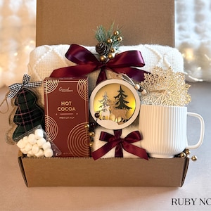 May include: A brown gift box filled with a cozy white blanket, a mug, a box of hot cocoa mix, marshmallows, and a decorative wooden ornament with a deer and a pine tree. The box is decorated with a burgundy ribbon and a gold snowflake.
