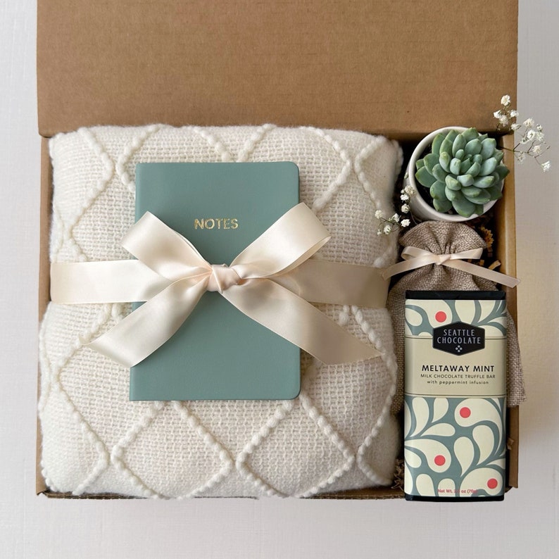 K&ouml;nnte beinhalten: Eine Geschenkbox gef&uuml;llt mit einer wei&szlig;en Wolldecke, einem t&uuml;rkisfarbenen Notizbuch mit dem Wort "NOTES" auf dem Einband, einer kleinen Sukkulente im Topf und einer Schokoladentafel mit der Aufschrift "Seattle Chocolate Meltaway Mint Milk Chocolate Truffle Bar with Peppermint Infusion" auf der Verpackung.