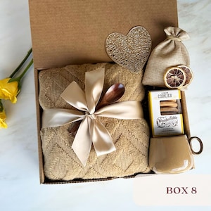May include: A gift box filled with a brown throw blanket, a wooden spoon, a bag of lemon cookies, a small bag of dried lemon slices, and a mug. The box is decorated with a heart-shaped cutout and a ribbon.
