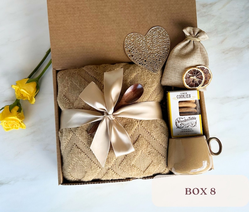 May include: A gift box filled with a brown throw blanket, a wooden spoon, a bag of lemon cookies, a small bag of dried lemon slices, and a mug. The box is decorated with a heart-shaped cutout and a ribbon.