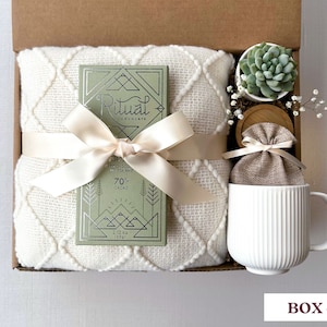 May include: A gift box filled with a white blanket, a bar of Ritual chocolate, a small potted succulent, a white mug, and a small burlap bag. The chocolate bar is wrapped in a green and silver foil with the text "Ritual Chocolate" and "70% Cacao".