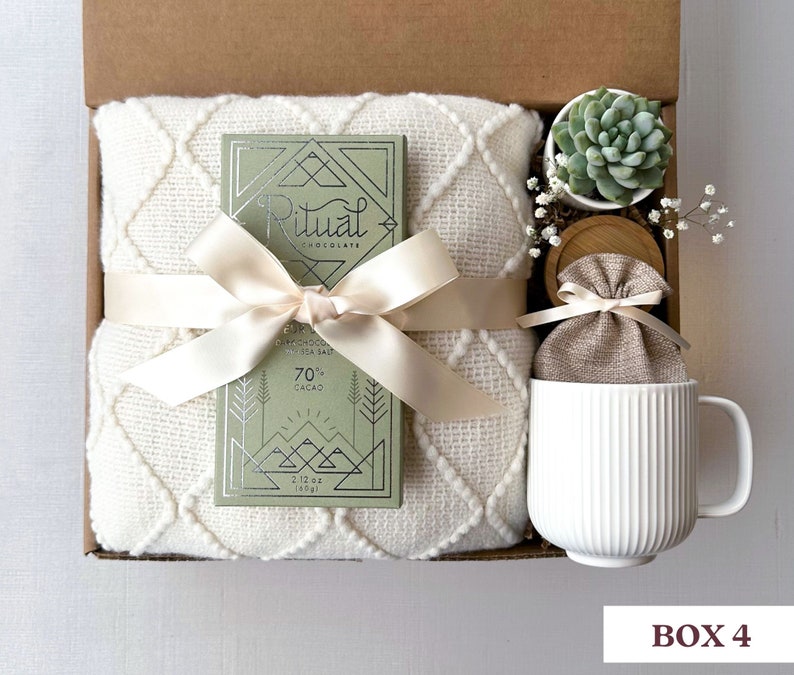May include: A gift box filled with a white blanket, a bar of Ritual chocolate, a small potted succulent, a white mug, and a small burlap bag. The chocolate bar is wrapped in a green and silver foil with the text "Ritual Chocolate" and "70% Cacao".