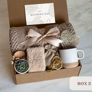 May include: A brown cardboard gift box filled with a variety of items, including a knitted throw blanket, a small potted succulent, a mug, a heart-shaped decorative object, a small bag, and a card that reads "Happy Mother's Day".