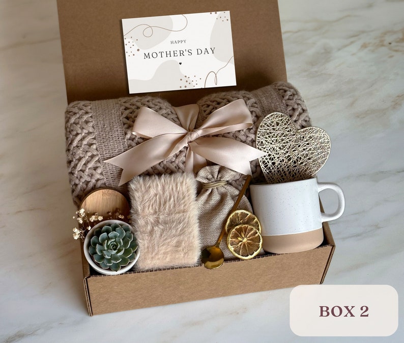 May include: A brown cardboard gift box filled with a variety of items, including a knitted throw blanket, a small potted succulent, a mug, a heart-shaped decorative object, a small bag, and a card that reads "Happy Mother's Day".