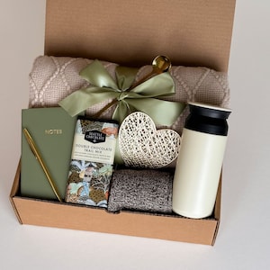 May include: A brown cardboard gift box filled with a variety of items, including a blanket, a notebook, a pen, a chocolate bar, a heart-shaped decorative object, and a white travel mug. The box is open and the items are visible.
