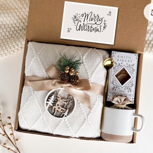 May include: A white gift box with a white blanket, a mug, a bag of mini gingerbread cookies, and a card that says "Merry Christmas".