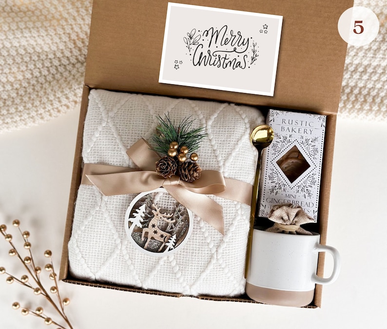 May include: A white gift box with a white blanket, a mug, a bag of mini gingerbread cookies, and a card that says "Merry Christmas".