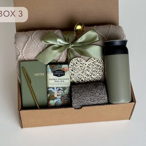 May include: A brown cardboard box with the text "BOX 3" on the front. The box is filled with a variety of items, including a blanket, a notebook, a pen, a chocolate bar, a heart-shaped decoration, and a green thermos.