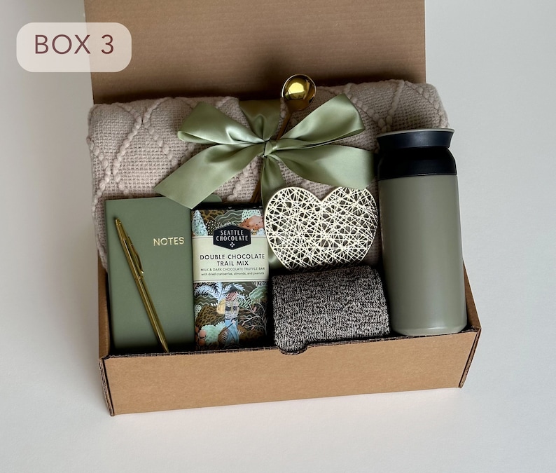 May include: A brown cardboard box with the text "BOX 3" on the front. The box is filled with a variety of items, including a blanket, a notebook, a pen, a chocolate bar, a heart-shaped decoration, and a green thermos.