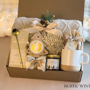 Puede incluir: Una caja de regalo r&uacute;stica de invierno llena de una taza blanca, chocolate, una cuchara dorada, una vela, un &aacute;rbol de madera decorativo, un copo de nieve dorado y una manta crema. La caja est&aacute; atada con una cinta dorada.