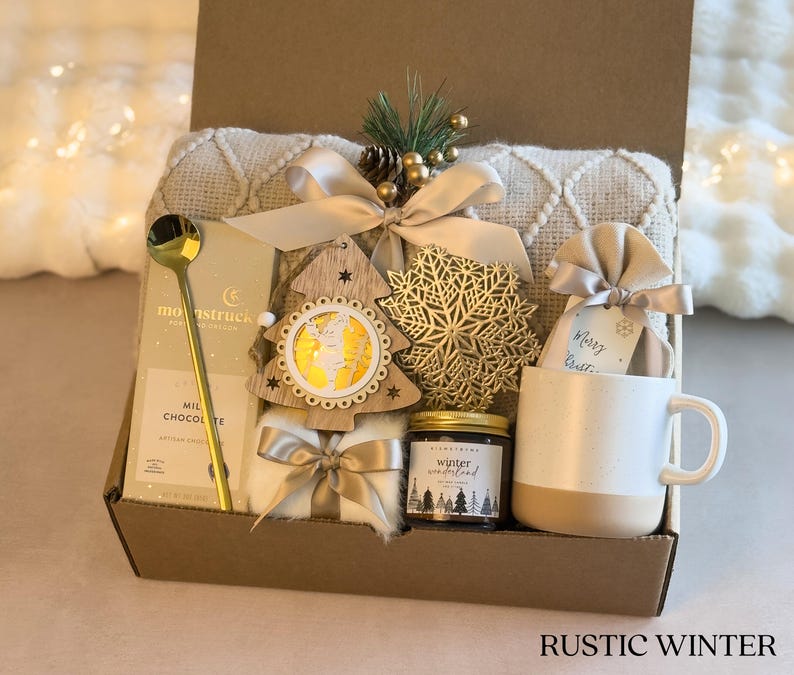 Puede incluir: Una caja de regalo r&uacute;stica de invierno llena de una taza blanca, chocolate, una cuchara dorada, una vela, un &aacute;rbol de madera decorativo, un copo de nieve dorado y una manta crema. La caja est&aacute; atada con una cinta dorada.