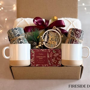 May include: A gift basket with a cozy theme, featuring two mugs, a pair of knitted socks, a box of chocolate chip cookies, and a wooden reindeer ornament. The basket is labeled "Fireside Duo".