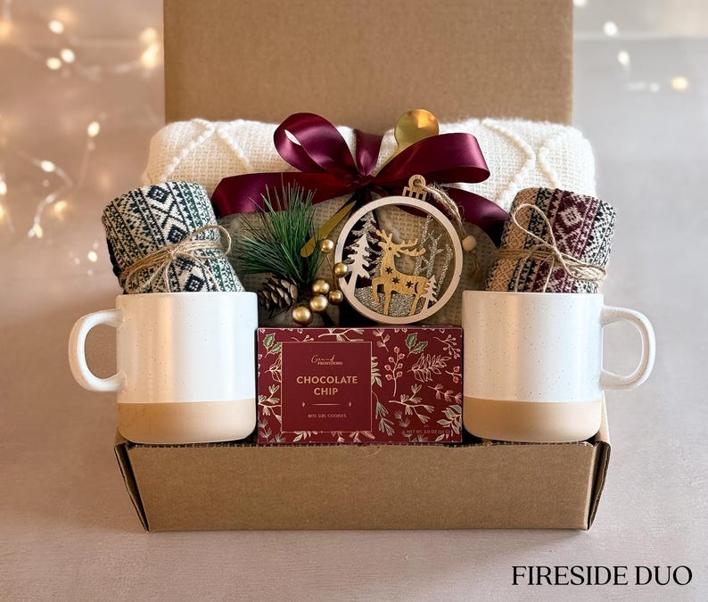 May include: A gift basket with a cozy theme, featuring two mugs, a pair of knitted socks, a box of chocolate chip cookies, and a wooden reindeer ornament. The basket is labeled "Fireside Duo".