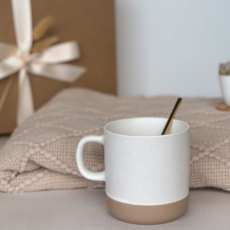 Puede incluir: Una taza de cer&aacute;mica blanca con dise&ntilde;o moteado y base marr&oacute;n. Una cuchara dorada descansa dentro de la taza. La taza est&aacute; frente a una manta beige y una caja de regalo envuelta con una cinta crema.