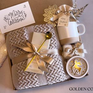 Puede incluir: Una caja de regalo navide&ntilde;a con una manta de punto beige, una taza blanca y beige, una cuchara dorada y un adorno de madera con un reno. La caja tambi&eacute;n contiene una tarjeta que dice "Merry Christmas!" y est&aacute; atada con una cinta dorada.