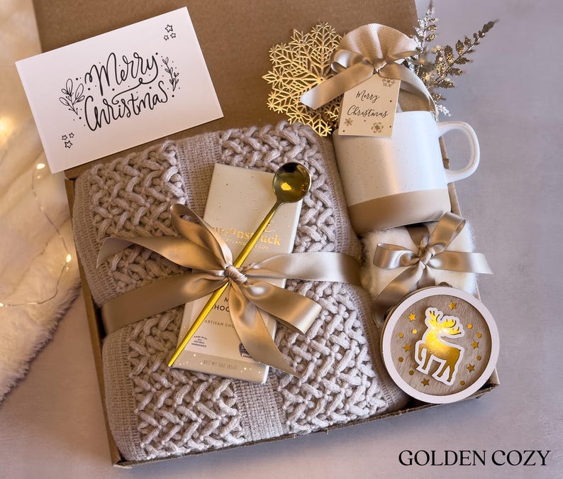 Puede incluir: Una caja de regalo navide&ntilde;a con una manta de punto beige, una taza blanca y beige, una cuchara dorada y un adorno de madera con un reno. La caja tambi&eacute;n contiene una tarjeta que dice "Merry Christmas!" y est&aacute; atada con una cinta dorada.
